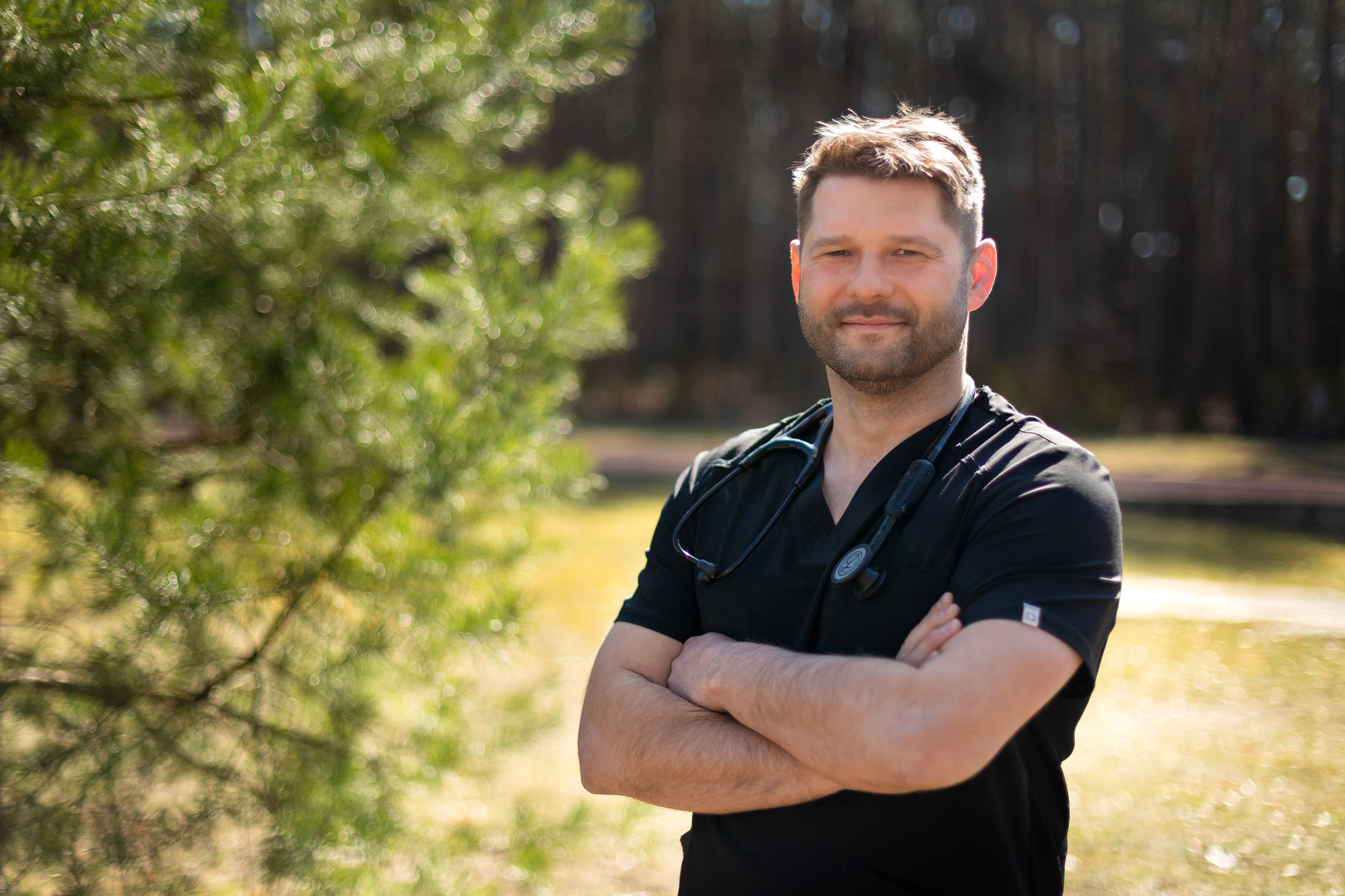 dr hab. Daniel Śliż ubrany w czarny strój medyczny, z założonym na szyję stetoskopem, stoi na zewnątrz w otoczeniu zieleni i drzew. Ma skrzyżowane ręce na piersiach. Tło jest rozmyte, ale widać trawę i drzewa.