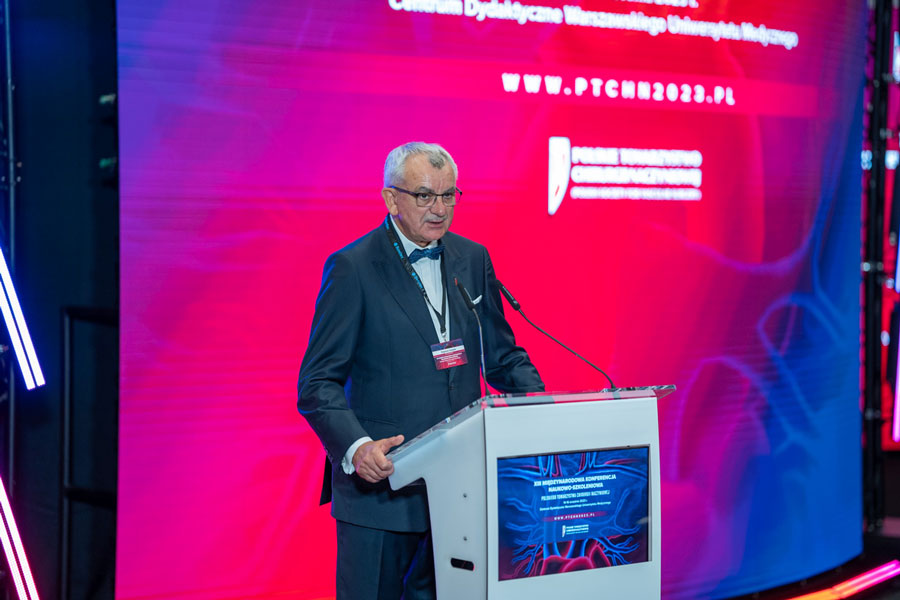 A smartly dressed man stands behind a lectern and speaks into a microphone. In the background behind him is a screen - a telescreen with the name of the conference.