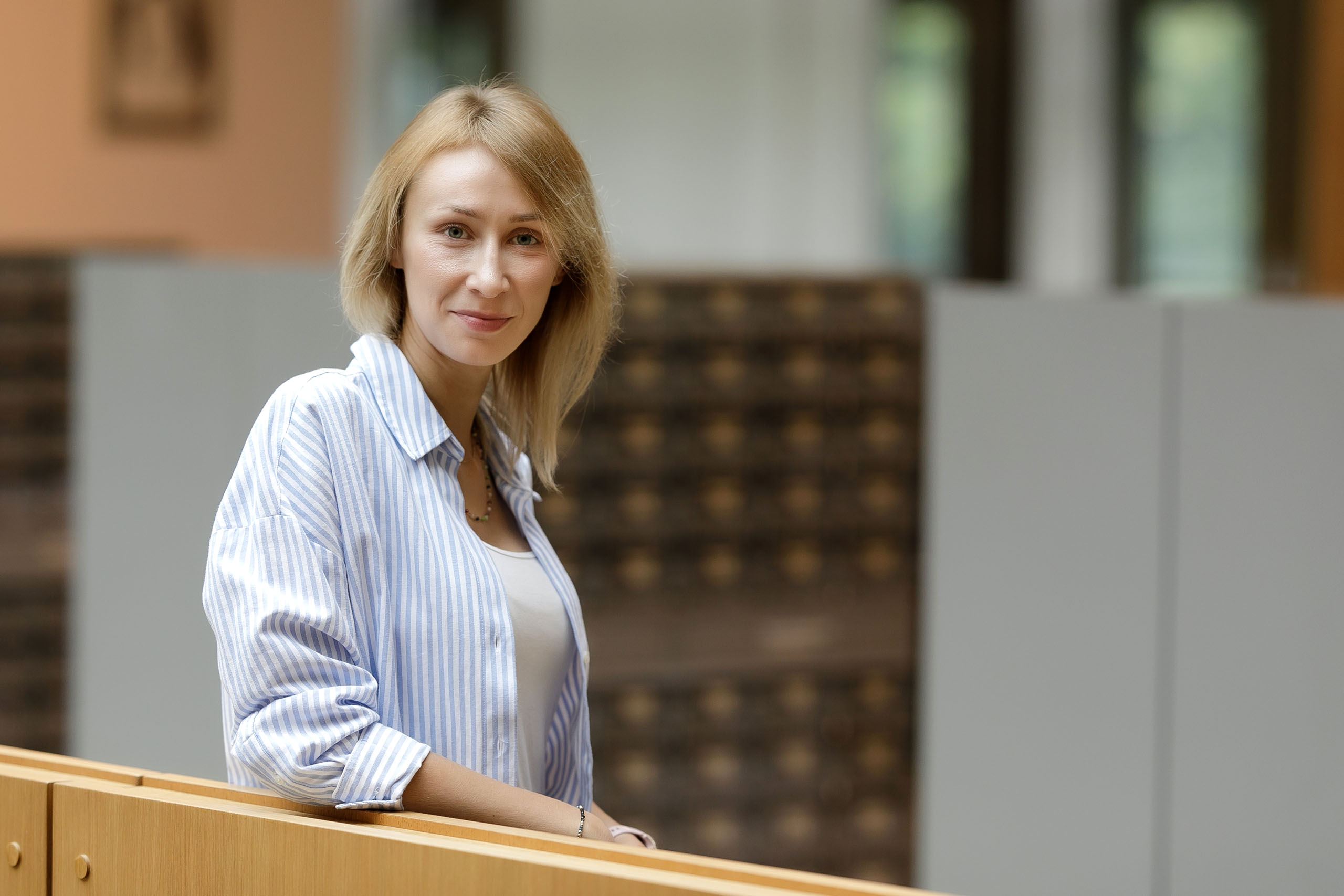 Młoda kobieta z jasnymi włosami opiera się o drewnianą balustradę. Uśmiecha się do aparatu. W tle regały biblioteczne.