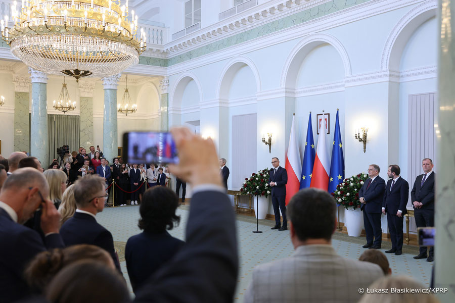 The ceremony takes place in a representative hall with high ceilings and a large crystal chandelier. The interior is decorated with columns and walls in white and green colors. In the foreground, a group of people can be seen standing with their backs to the camera, some of whom are photographing or recording the event with their smartphones. On the right side of the frame, a man is speaking from a podium, next to which are four flags—two Polish and two European Union flags—and several men in suits. Transl