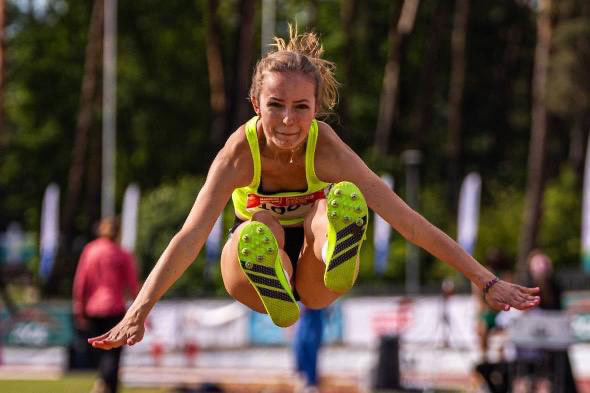 A female athlete in bright green sportswear performs a long jump in an athletics stadium. She is captured in the air with her arms outstretched and legs bent. Blurred silhouettes of people and trees are visible in the background.