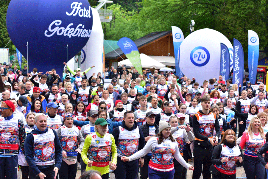 Duża grupa ludzi uczestniczących w wydarzeniu na świeżym powietrzu - Biegu po Nowe Życie. Uczestnicy mają na sobie kolorowe koszulki i numery startowe. W tle widoczne są dwa duże balony reklamowe. W tle znajdują się również drzewa i drewniane budynki.