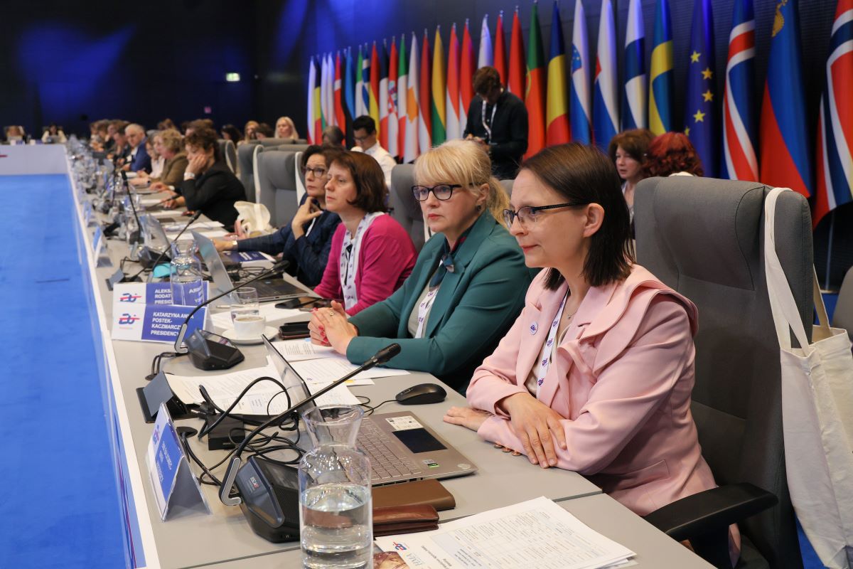 A long table in a conference room. Seated at it are about 20 people. In the foreground is a middle-aged woman wearing a light pink jacket. Each person has a microphone, laptop and documents in front of him. Behind the participants is a row of flags of various countries and a flag of the European Union. The atmosphere is formal and professional.