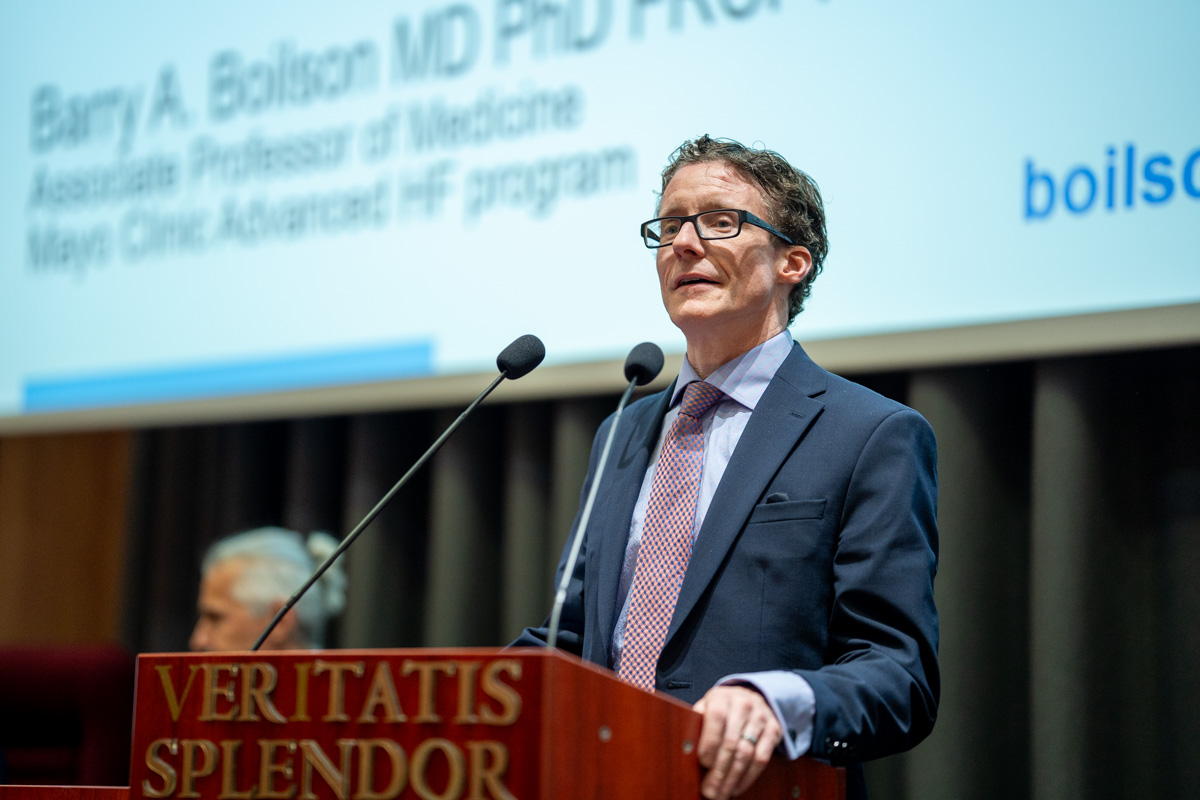 Mężczyzna w średnim wieku, w okularach. Ma na sobie ciemny garnitur, jasnoniebieską koszulę i jasnoróżowy krawat. Stoi za mównicą i przemawia. Mównica jest brązowa i ma napis "VERITATIS SPLENDOR" w kolorze złotym. W tle znajduje się ekran multimedialny z tekstem.