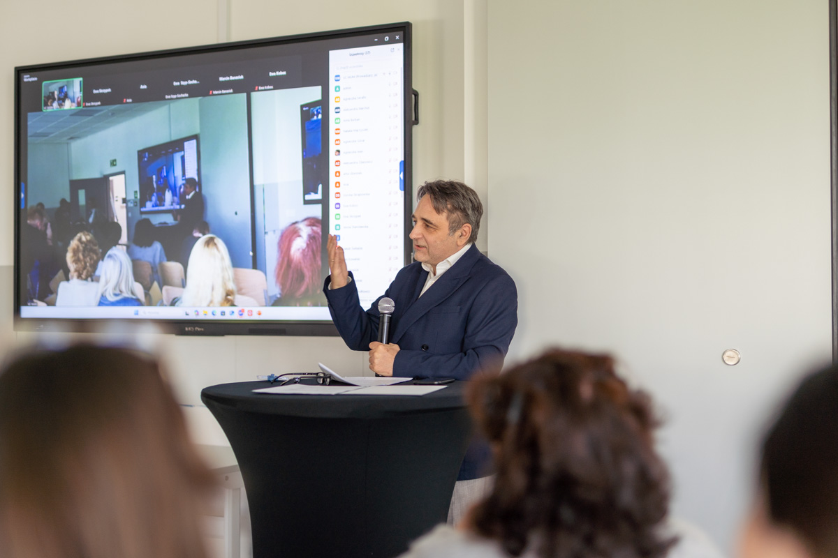 "Mężczyzna - dr hab. Piotr Dziechciarz - w garniturze stoi za czarną mównicą, trzyma mikrofon i gestykuluje ręką. Za nim znajduje się duży ekran wyświetlający wideokonferencję z wieloma uczestnikami widocznymi na ekranie. Sala jest przygotowana do prezentacji lub wykładu, z kilkoma osobami siedzącymi i zwróconymi w stronę mówcy.