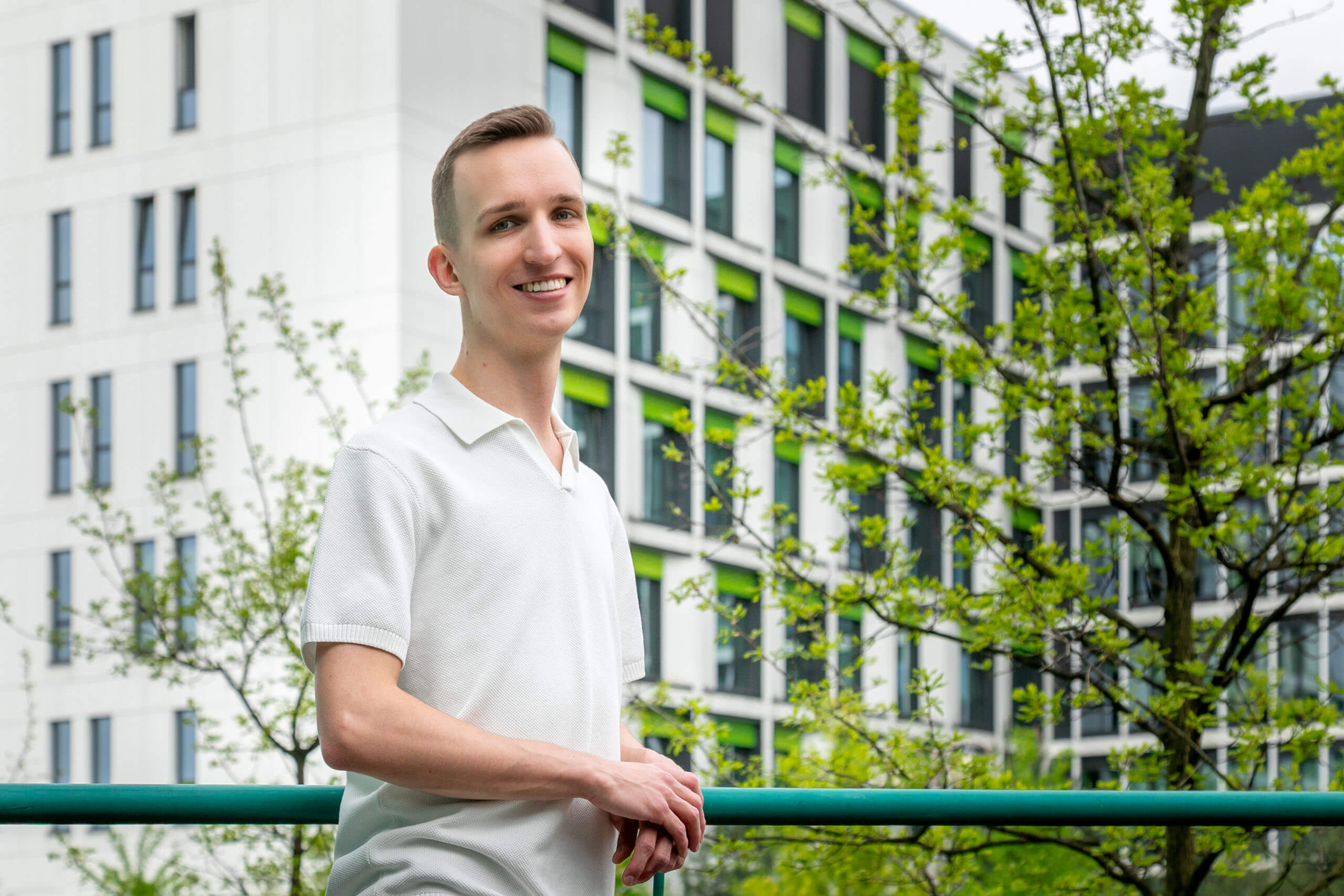 Młody mężczyzna, patrzy przed siebie i uśmiecha się do zdjęcia. Ma na sobie białą koszulkę polo i jasnoszare spodnie. Tło stanowi nowoczesny budynek z dużymi oknami oraz drzewa z zielonymi liśćmi. 