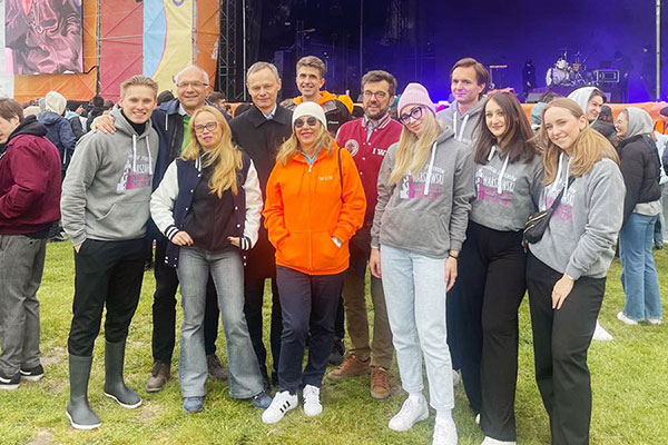 Eleven people standing on the grass. In the background is a stage with musical instruments visible and a crowd of participants. The participants are dressed in a variety of outfits - from jackets to sweatshirts and pants. Several people are wearing gray sweatshirts with graphics depicting the Palace of Culture and Science. A colorful advertising banner is visible on the left side of the frame.