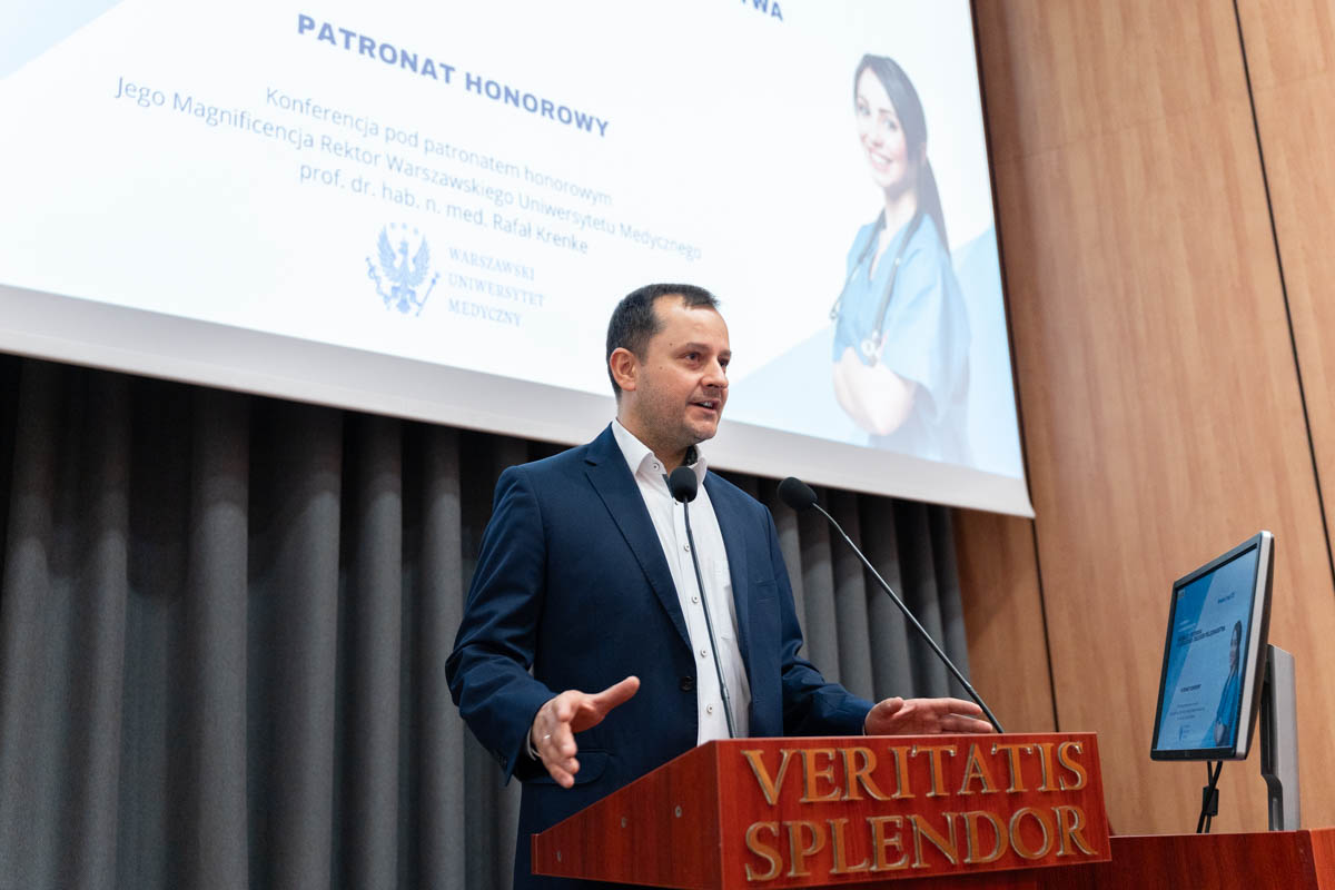 Na pierwszym planie mężczyzna w garniturze, który przemawia na konferencji. Stoi za mównicą z napisem 'VERITATIS SPLENDOR' i mówi do mikrofonu. W tle znajduje się ekran projekcyjny z tekstem: 'PATRONAT HONOROWY Konferencja pod patronatem honorowym Jego Magnificencja Rektor Warszawskiego Uniwersytetu Medycznego. Na ekranie widoczna jest również kobieta w niebieskim fartuchu medycznym, uśmiechająca się i trzymająca ręce skrzyżowane na piersi.