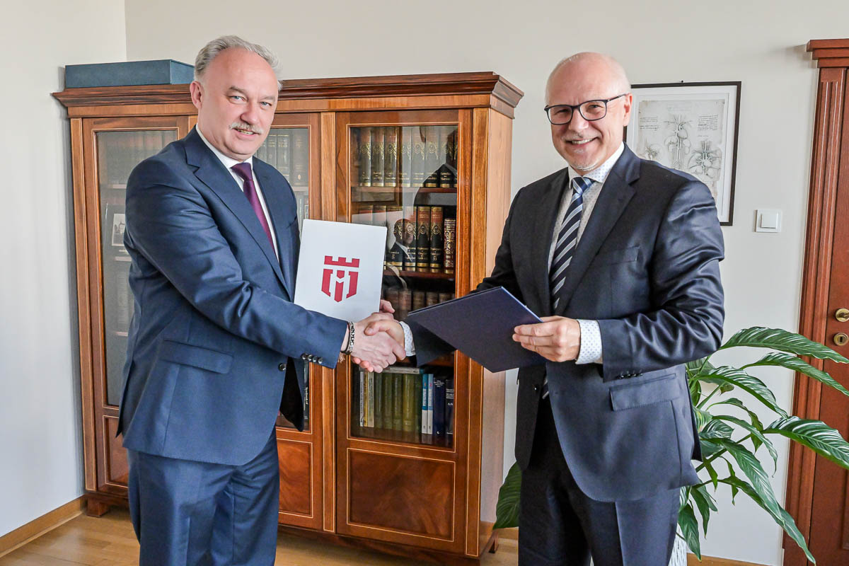 Rektorzy obu uczelni. Dwóch mężczyzn w ciemnych garniturach podaje sobie ręce. Mężczyzna po lewej trzyma czarną teczkę. Na teczce widnieje czerwone logo na białym tle. W tle znajduje się drewniana szafa z książkami. Po prawej stronie zdjęcia stoi zielona roślina doniczkowa.