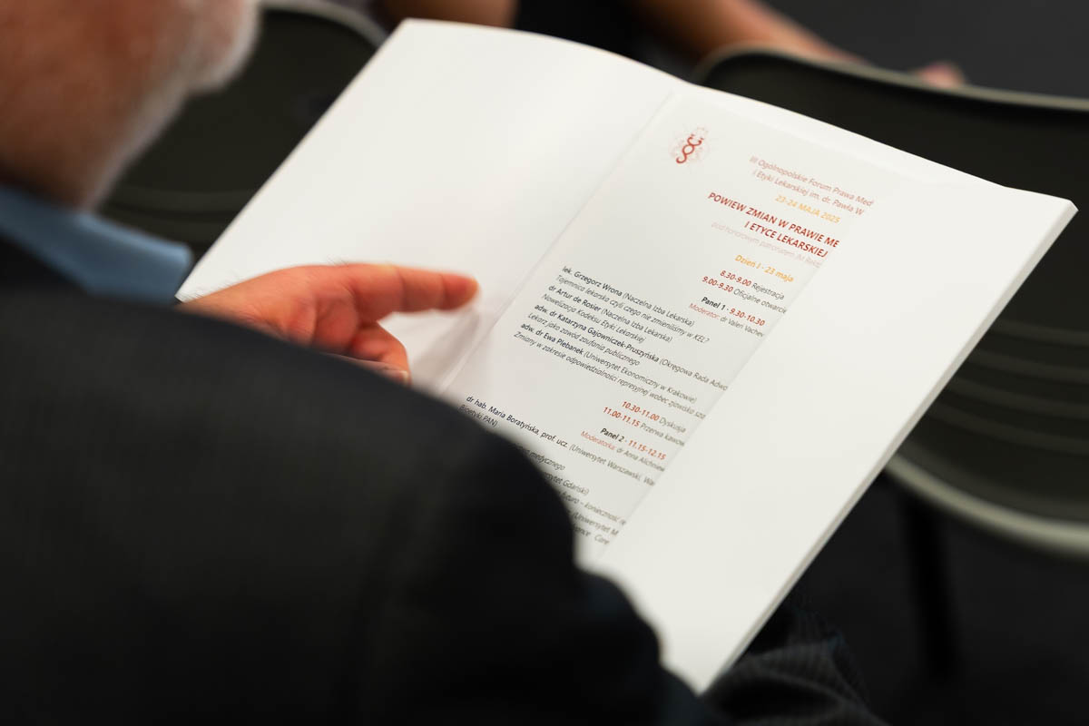 A person holds open a conference brochure with the program of the 3rd National Forum on Medical Law and Ethics. The headings, names of speakers and panel times are visible. A finger points to a section of text. In the background are fuzzy chairs.