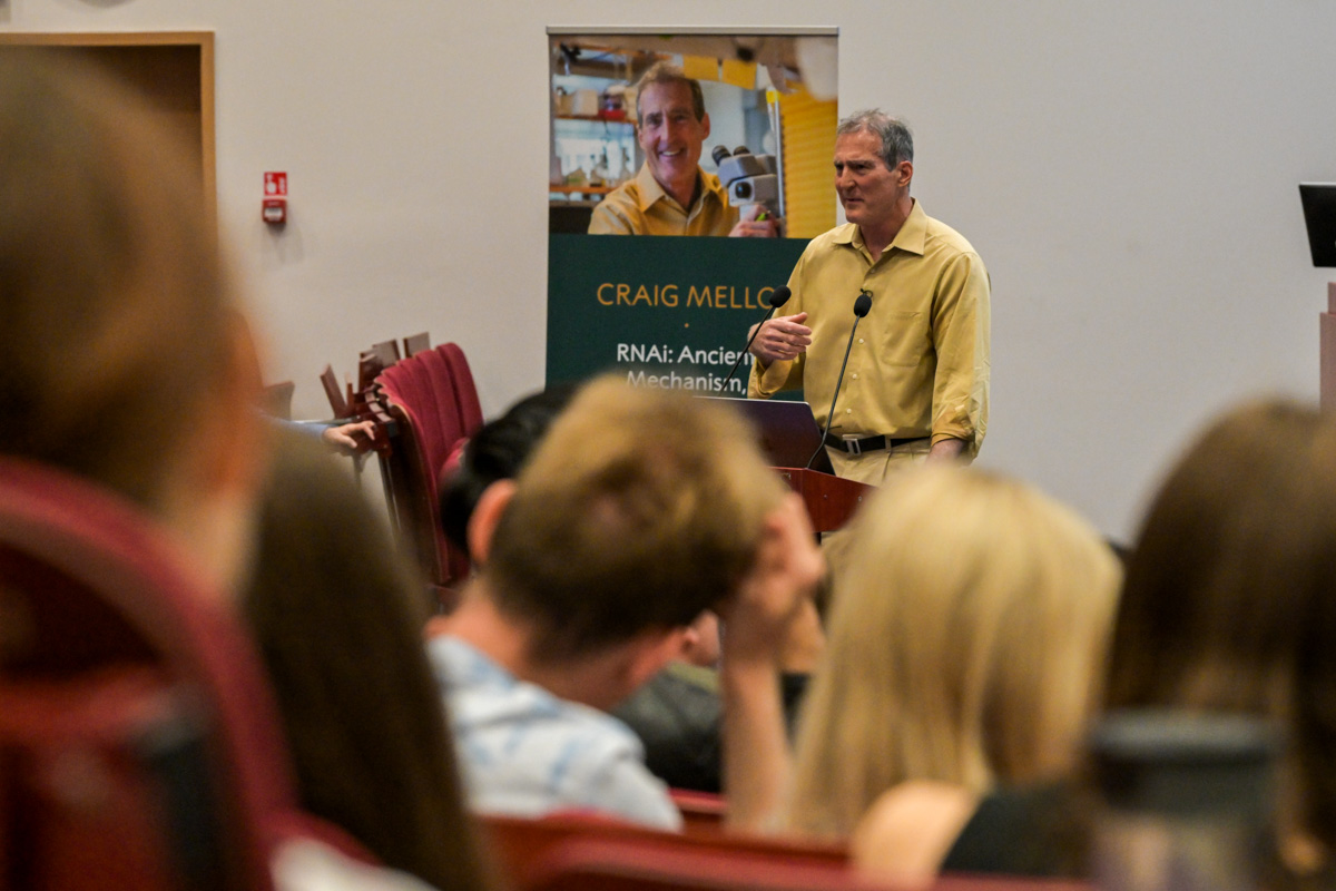 Prof. Craig C. Mello, man in a yellow shirt with a microphone stands in front of an audience in a lecture hall. He gestures with his hand. A poster in the background reads 'CRAIG MELLO RNA: Ancient Mechanisms.