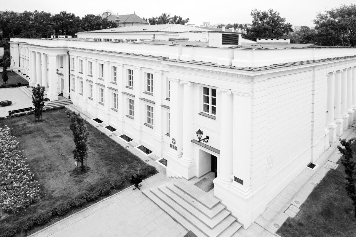 Czarno-białe zdjęcie przedstawia klasyczny, dwupiętrowy budynek na Kampusie Głównym Uniwersytetu Warszawskiego. Budynek otoczony jest trawnikiem i alejkami. Na pierwszym planie widoczne są schody prowadzące do głównego wejścia po prawej stronie. W tle znajdują się drzewa i inne budynki. To miejsce było świadkiem makabrycznej zbrodni.