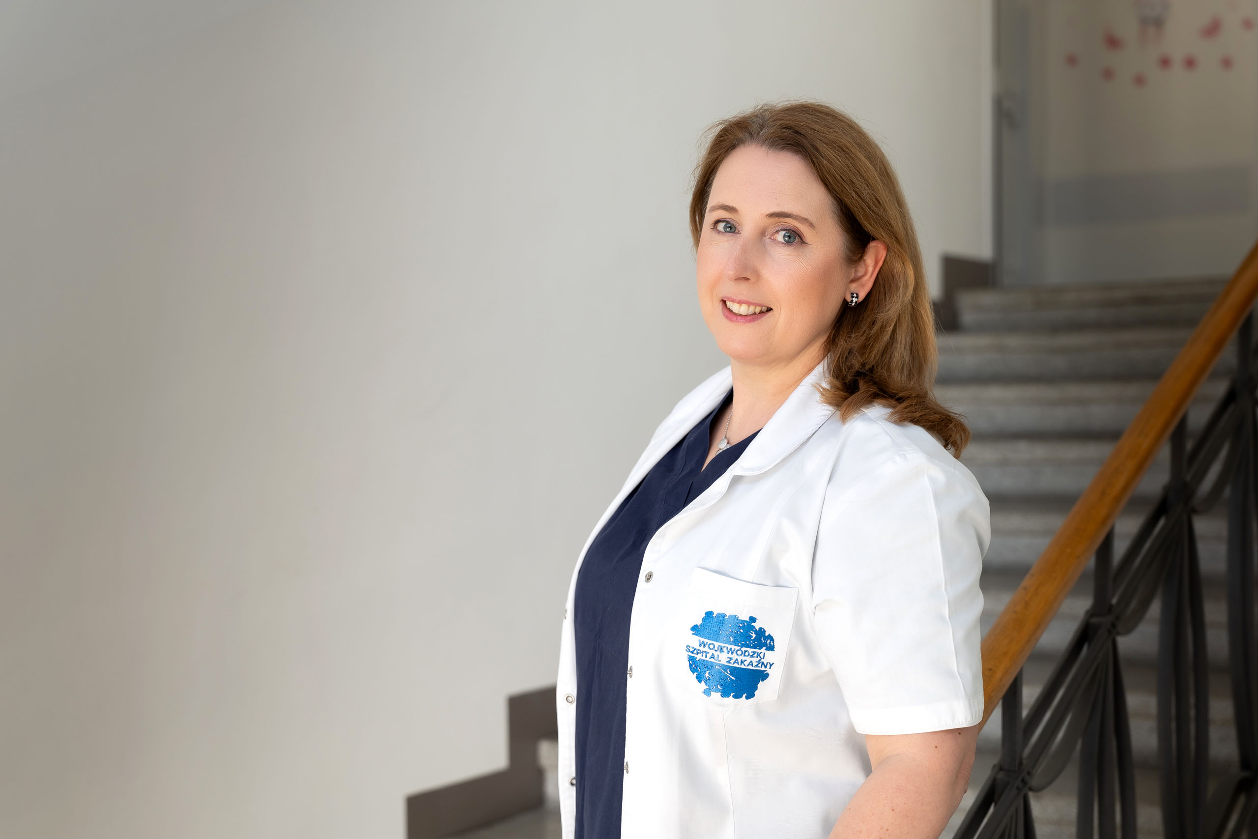 Kobieta stoi wewnątrz budynku obok schodów. Ma na sobie biały fartuch z emblematem po lewej stronie, który zawiera tekst i niebieski okrągły wzór. Tło zdjęcia pokazuje część schodów z metalowymi poręczami i stopniami prowadzącymi w górę.