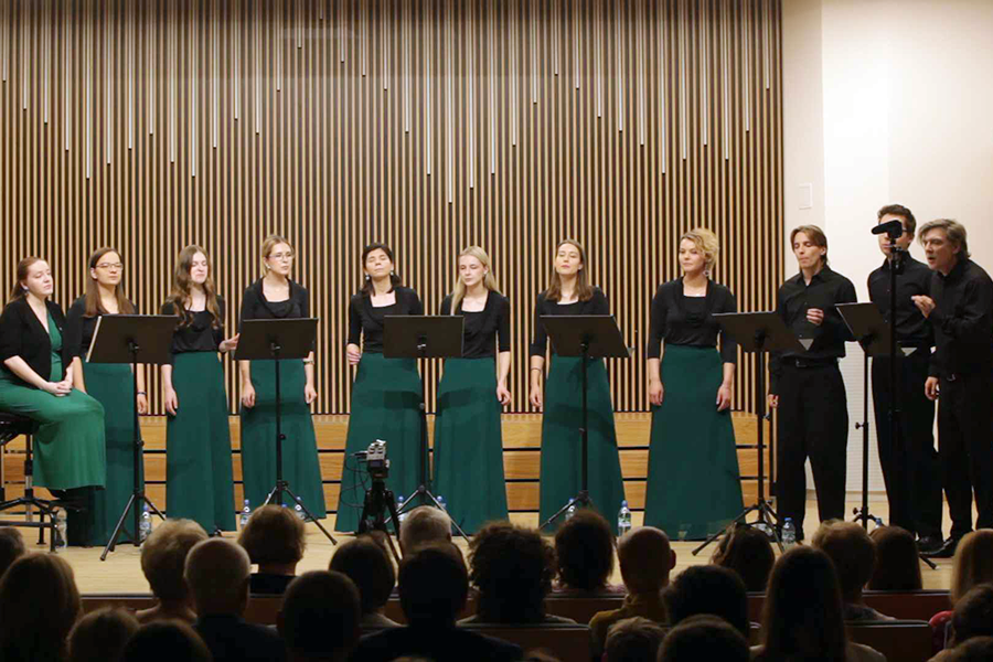 Dziesięć osób stoi na scenie w sali koncertowej, przed pulpitami z nutami. Siedem kobiet w zielonych spódnicach i czarnych bluzkach, trzech mężczyzn ubranych na czarno. W tle drewniana ściana akustyczna z pionowymi elementami. Na pierwszym planie widoczna publiczność siedząca na krzesłach.