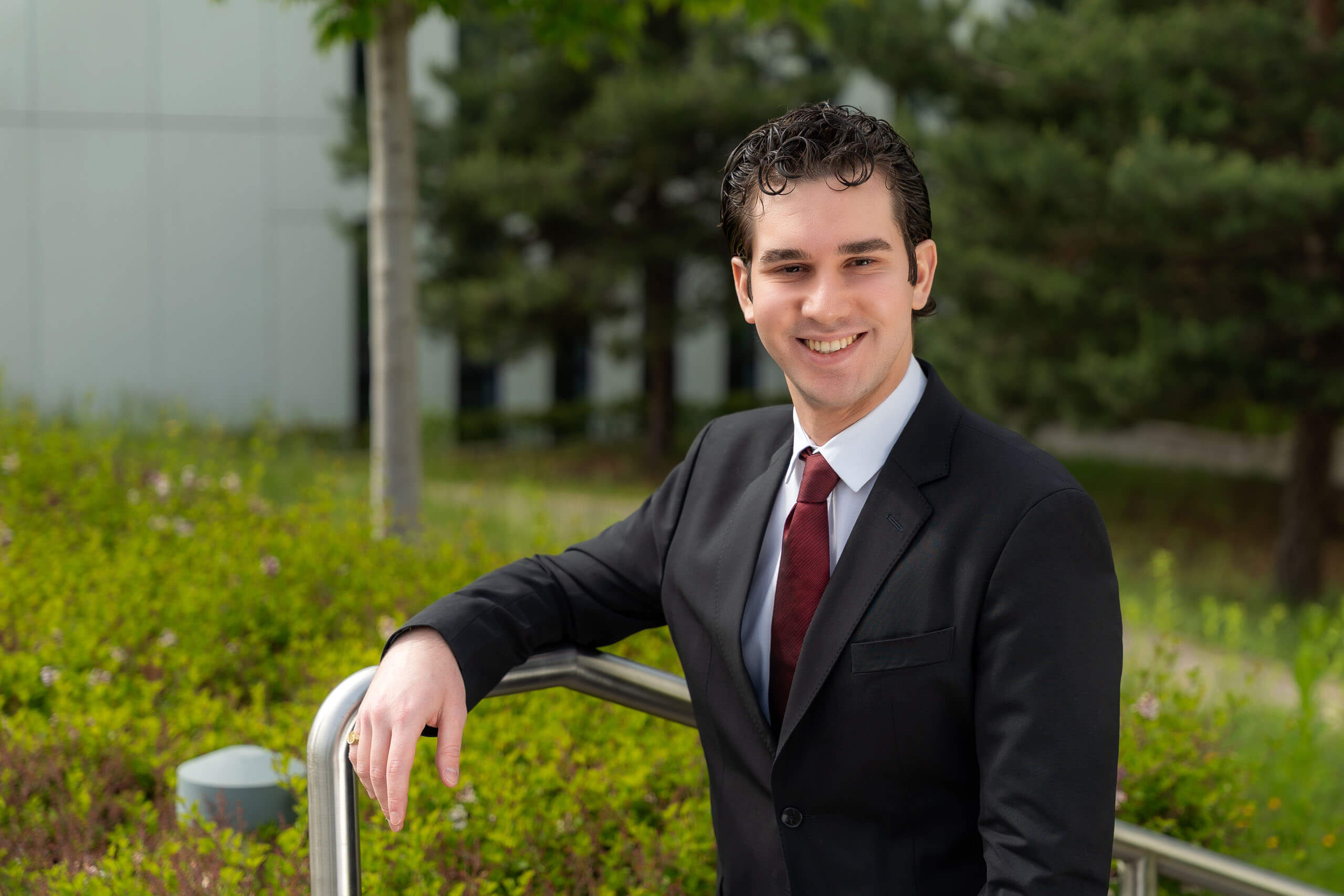 Młody mężczyzna w czarnej marynarce, białej koszuli i bordowym krawacie stoi na zewnątrz, oparty o metalową barierkę. W tle widoczne są zielone krzewy, drzewa i fragment budynku. Mężczyzna uśmiecha się. Ma ciemne włosy i ciemne oczy.
