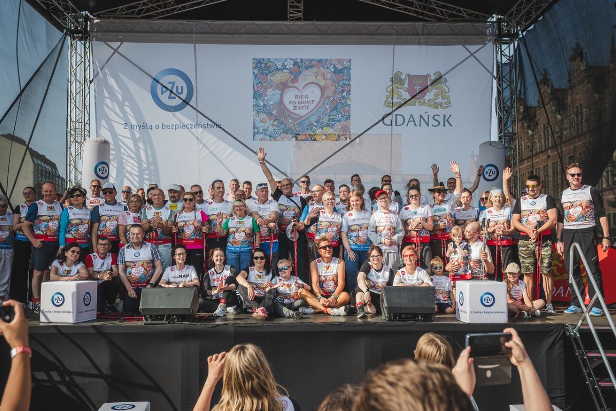 Grupa uczestników wydarzenia sportowego stojąca na scenie ubrana w koszulki z numerami startowymi. Część osób trzyma dyplomy lub medale, inni machają do zdjęcia. 