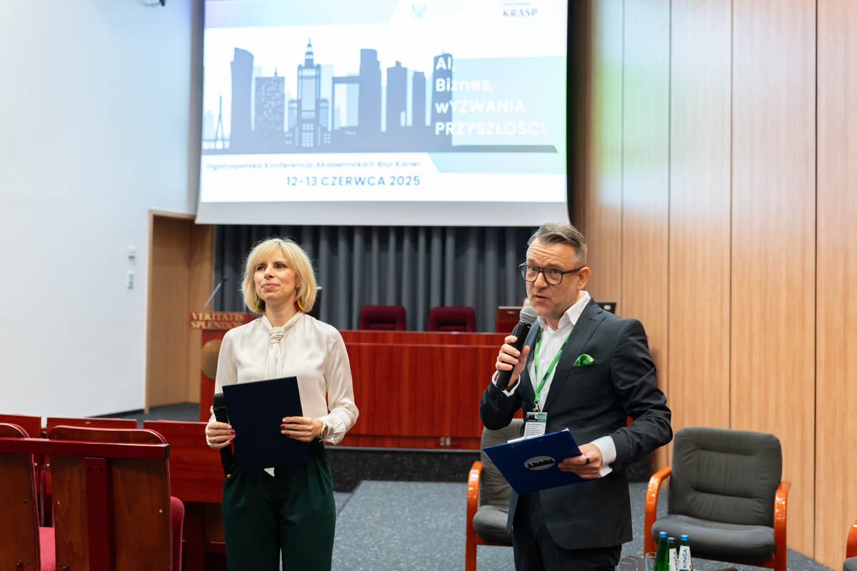 The two presenters stand on the conference stage. A woman in a white blouse and green trousers is holding a black briefcase, a man in a dark suit with a green pillowcase is holding a microphone and a blue briefcase.