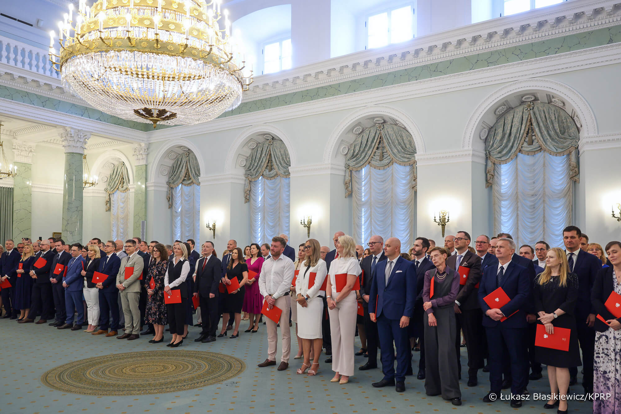 Grupa osób stoi w eleganckiej sali z wysokim sufitem, kolumnami i dużym żyrandolem. Wszyscy ubrani są formalnie i trzymają czerwone teczki. Ściany są jasne, okna duże z zielonymi zasłonami. Na podłodze leży zielony dywan.