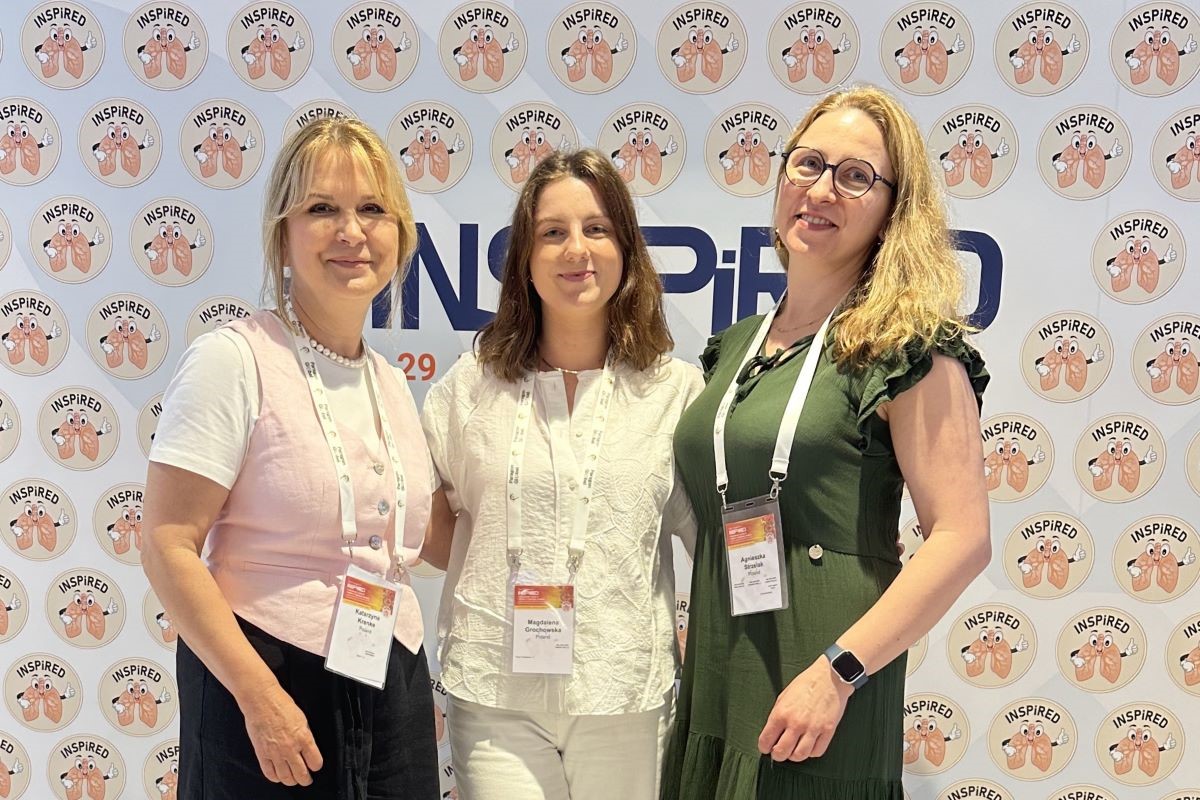 Three women standing in front of a wall with the ‘INSPIRED’ logo and a design depicting two hands holding a heart. Each of the women has an ID tag hanging around her neck.
