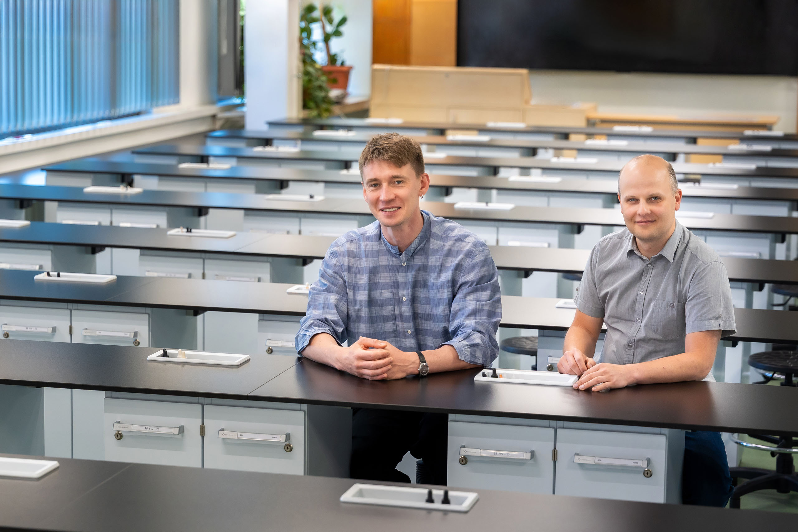 Dwóch mężczyzn siedzi w jasnej, nowoczesnej sali szkoleniowej. Wnętrze wyposażone jest w rzędy biurek z czarnymi blatami i białymi szufladami