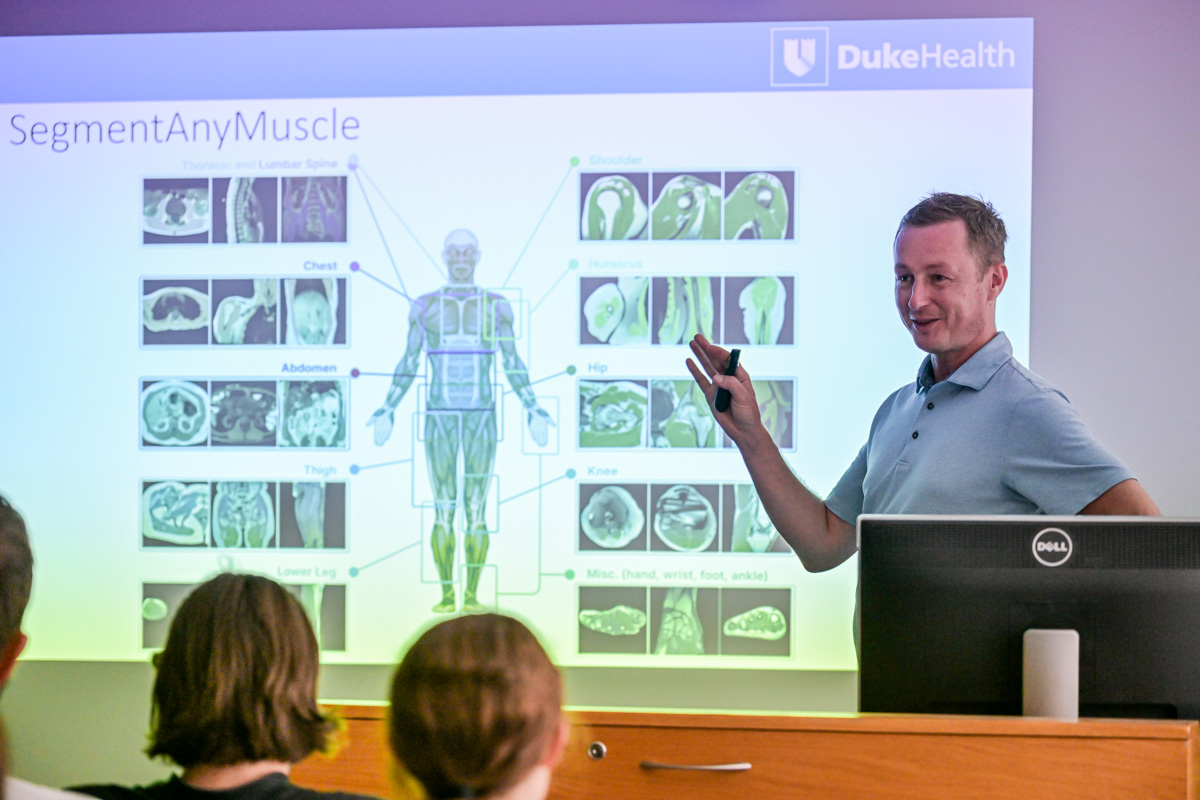 Prezentacja medyczna w sali wykładowej. Na ekranie slajd z tytułem „SegmentAnyMuscle” i logo DukeHealth. Widać kolorową ilustrację mięśni człowieka oraz zdjęcia MRI szyi, klatki piersiowej, brzucha, uda, kolana i stopy. Prezenter wskazuje slajd. W tle siedzą słuchacze, widoczni od tyłu.