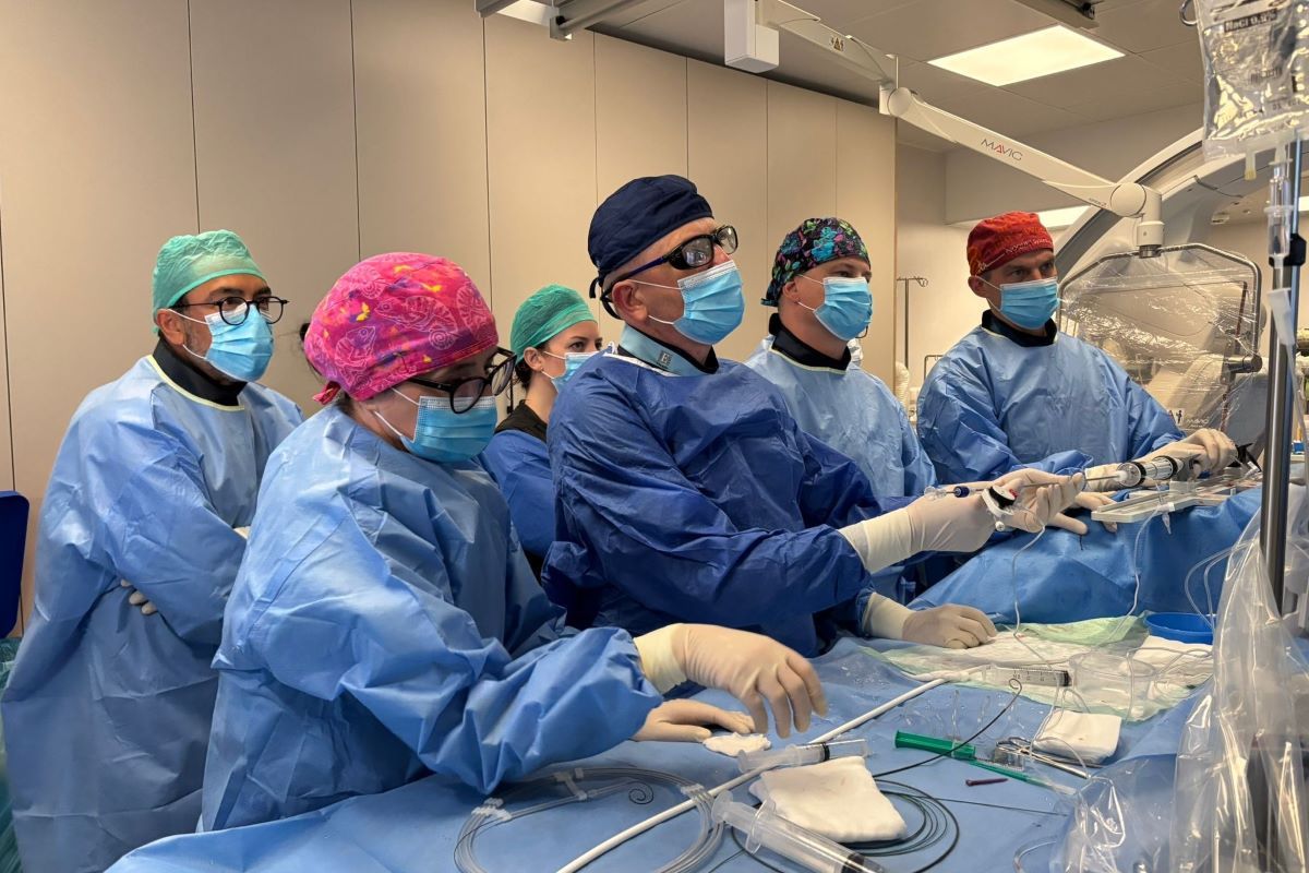 A group of men and women dressed in blue surgical gowns, caps, and gloves perform a medical procedure at an operating table, holding surgical instruments in their hands. In the background are operating lights and operating room monitors.