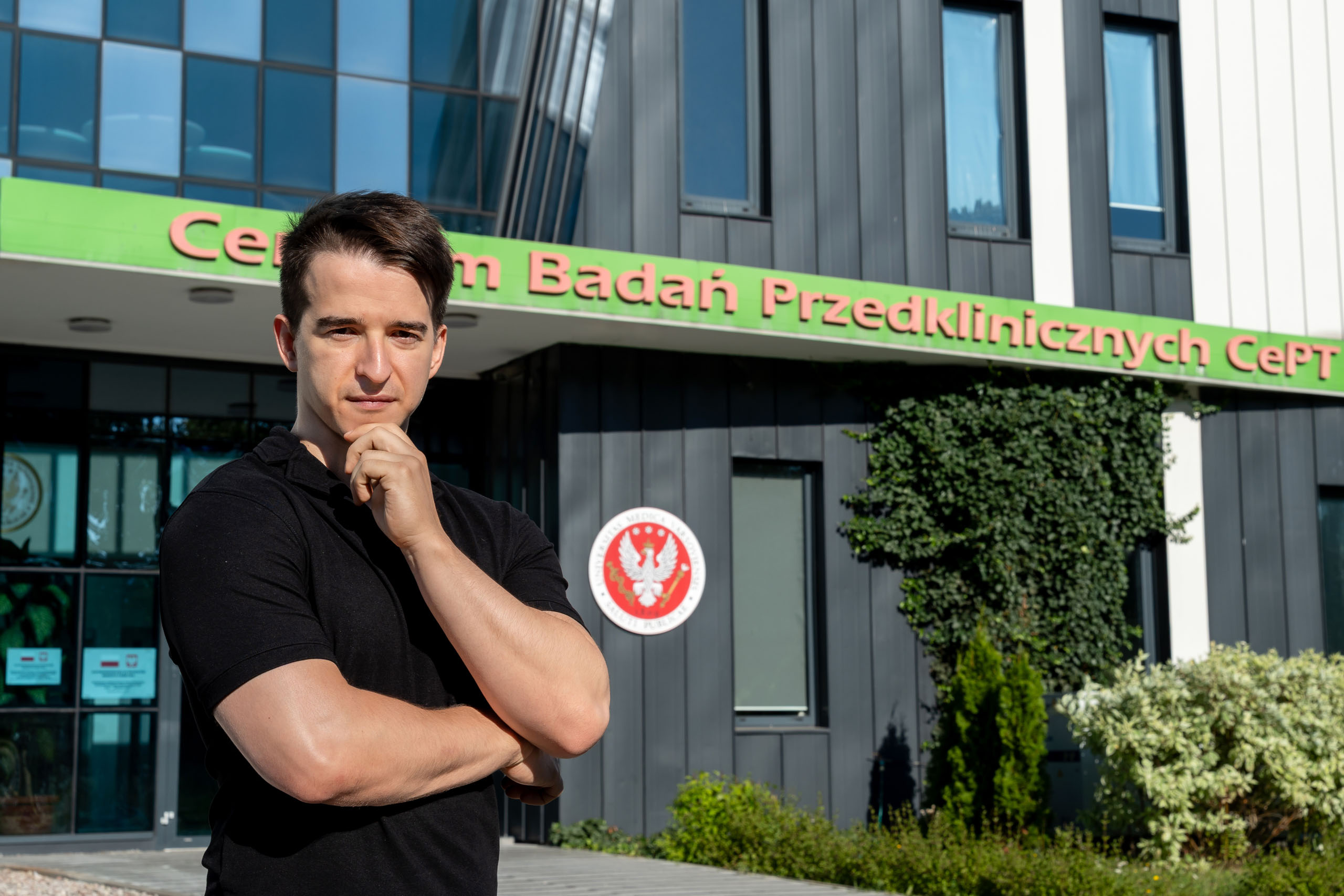 A man in a black T-shirt stands in front of the CePT Preclinical Research Center building. The building has gray walls and large windows. Above the entrance is a green sign with the name. Next to the door is a red coat of arms with a white eagle. In the background are decorative plants and shrubs.