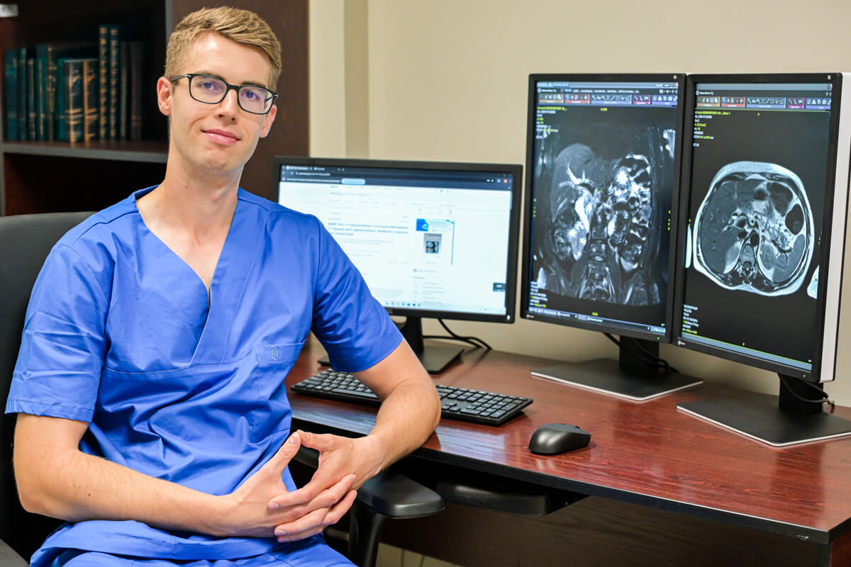 Młody mężczyzna, dr Krzysztof Bartnik, w okularach i niebieskim fartuchu lekarskim. Siedzi przy stole. Na stole stoją trzy ekrany komputerowe. Na dwóch z nich widać obrazy radiologiczne.