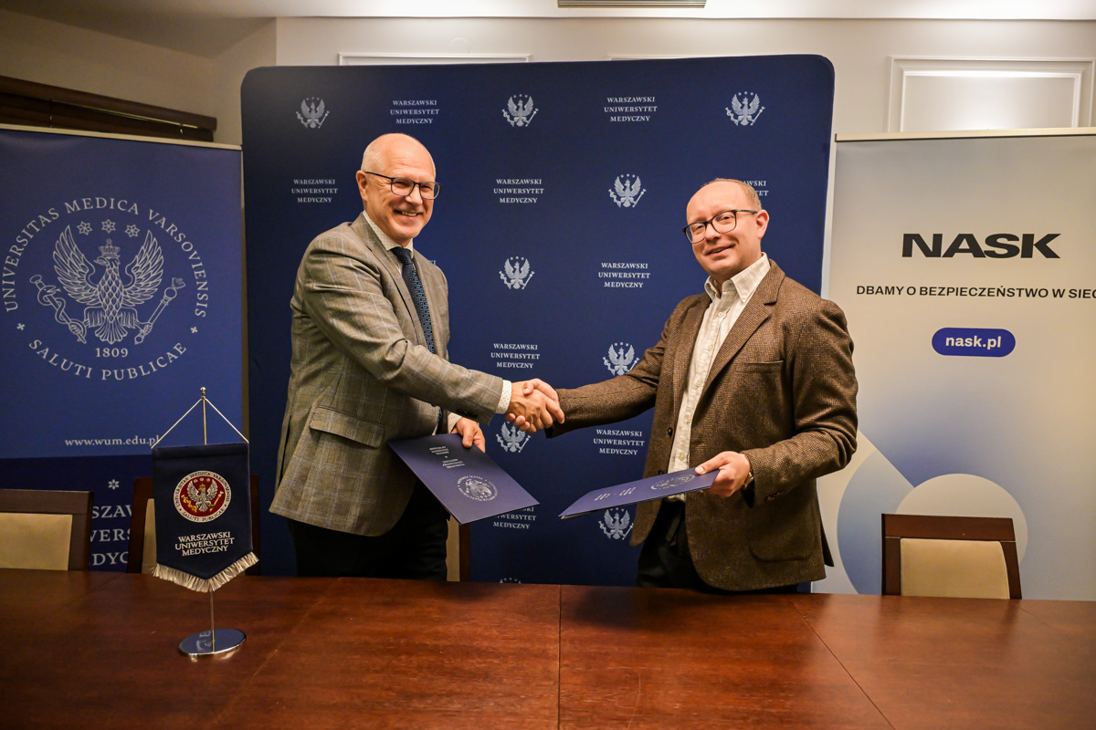 Dwóch mężczyzn ubranych elegancko ściska sobie dłonie. W drugiej ręce każdy trzyma teczkę z dokumentami. Uśmiechają się do obiektywu. W tle banery WUM i NASK