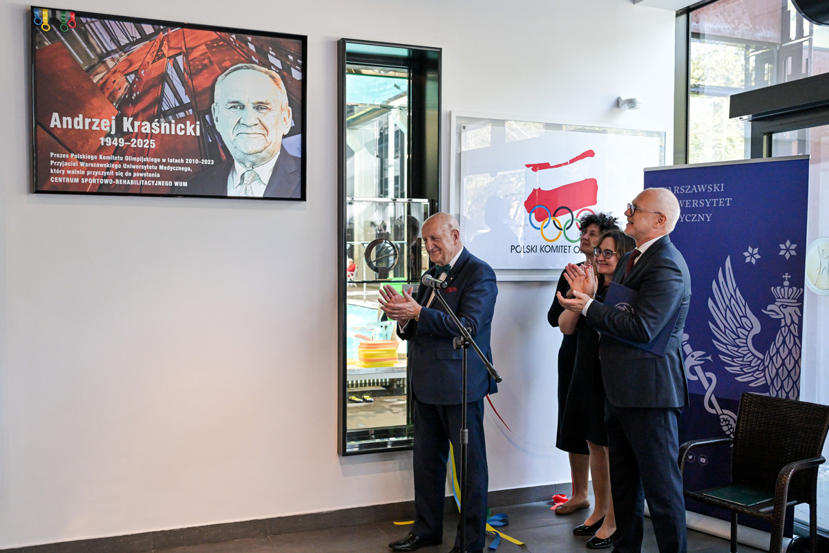 Trzy osoby stoją przy mikrofonie i klaszczą podczas odsłonięcia tablicy pamiątkowej Andrzeja Kraśnickiego (1949–2025). Tablica z portretem i tekstem znajduje się na ścianie po lewej. W tle witryna z trofeum Międzynarodowego Komitetu Olimpijskiego.