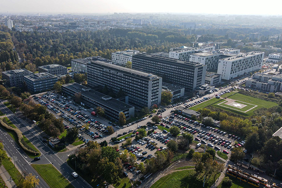 Zdjęcie kampusu z lotu ptaka. W centrum dwa duże, prostokątne budynki otoczone mniejszymi obiektami. Wokół widoczne parkingi z wieloma samochodami. Tereny zielone otaczają zabudowania. W tle rozciąga się panorama miasta aż po horyzont.