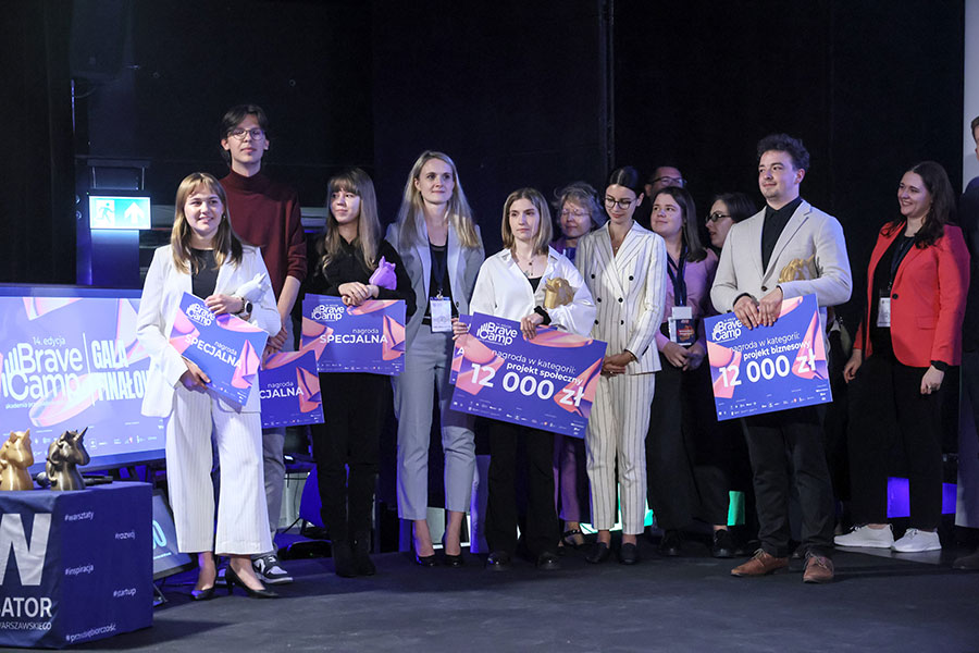 A group of young people, elegantly dressed, are standing on stage. Some of them are holding large cardboard checks in their hands. They are all smiling.