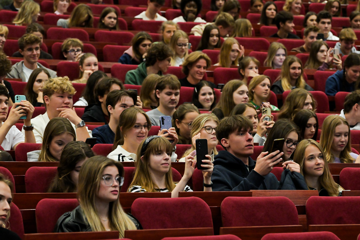 Grupa młodych kobiet i mężczyzn siedzi w audytorium, zwrócona w stronę sceny. Wiele osób trzyma telefony komórkowe, robiąc zdjęcia lub nagrywając filmy