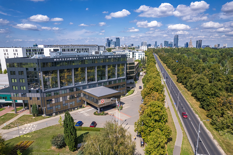Na pierwszym planie nowoczesny budynek z dużymi oknami i napisem „Warszawski Uniwersytet Medyczny” na dachu. Przed budynkiem znajdują się flagi. Po prawej stronie biegnie szeroka ulica otoczona drzewami. W tle panorama miasta z wieżowcami i biurowcami.