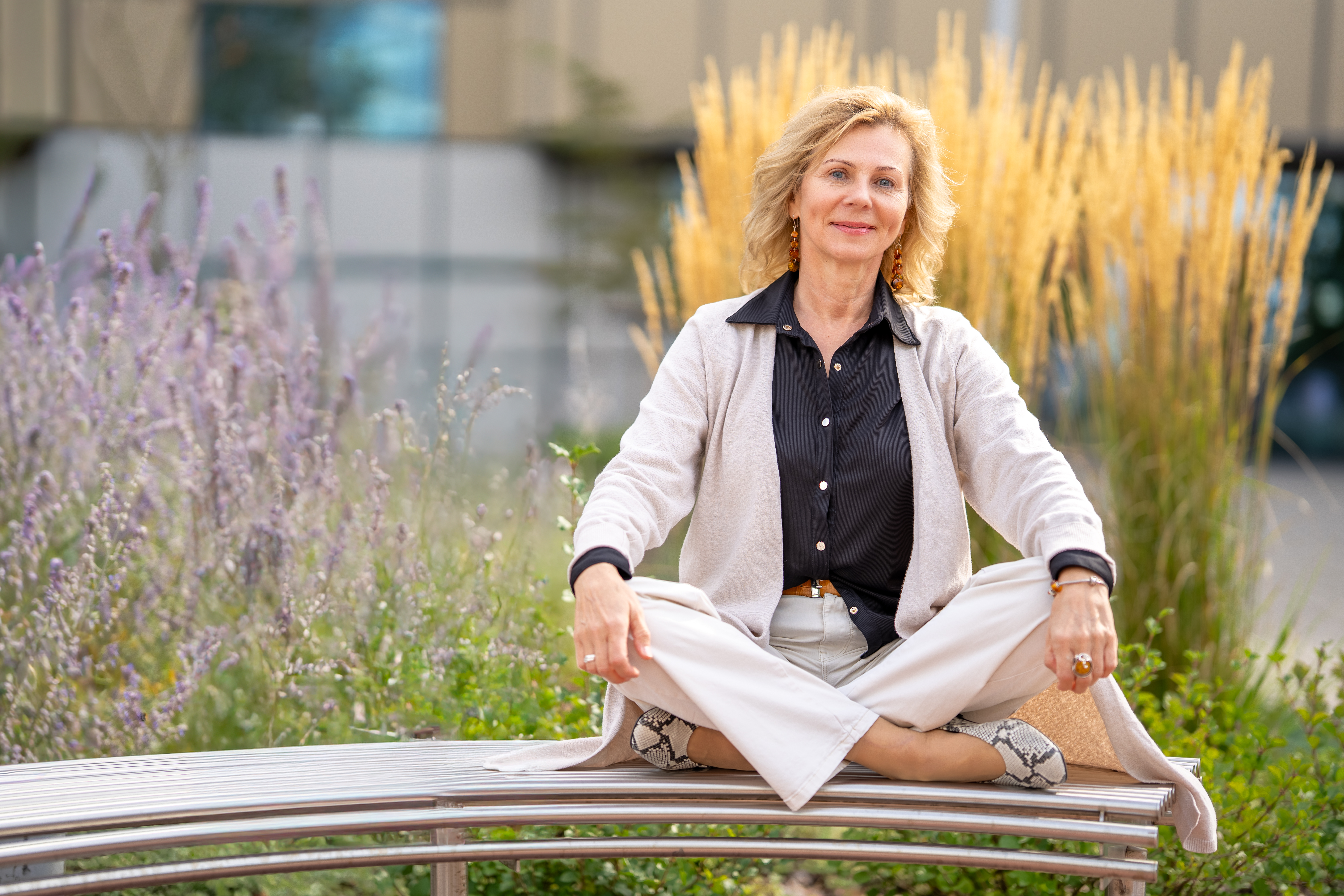Kobieta siedzi na ławce w ogrodzie, mając skrzyżowane nogi. Ubrana jest w jasne spodnie, czarną koszulę i jasny sweter. W tle widoczne są rośliny ozdobne.