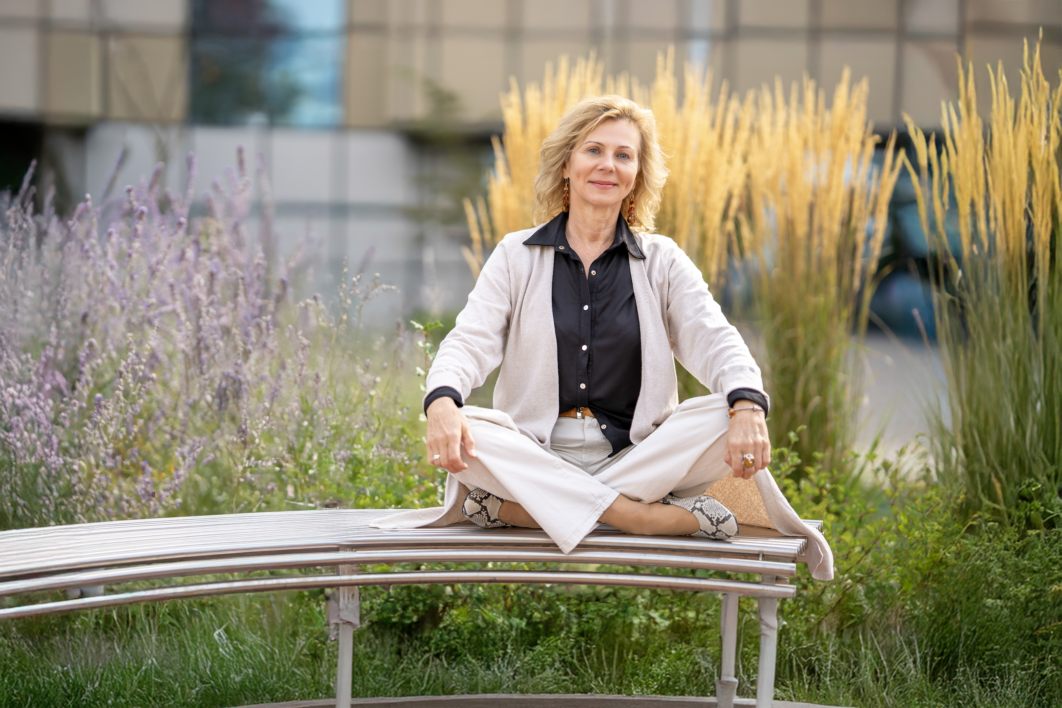 A woman, Prof. Dorota Włodarczyk, is sitting on a bench in the lotus position. She is smiling at the camera. She is surrounded by greenery.