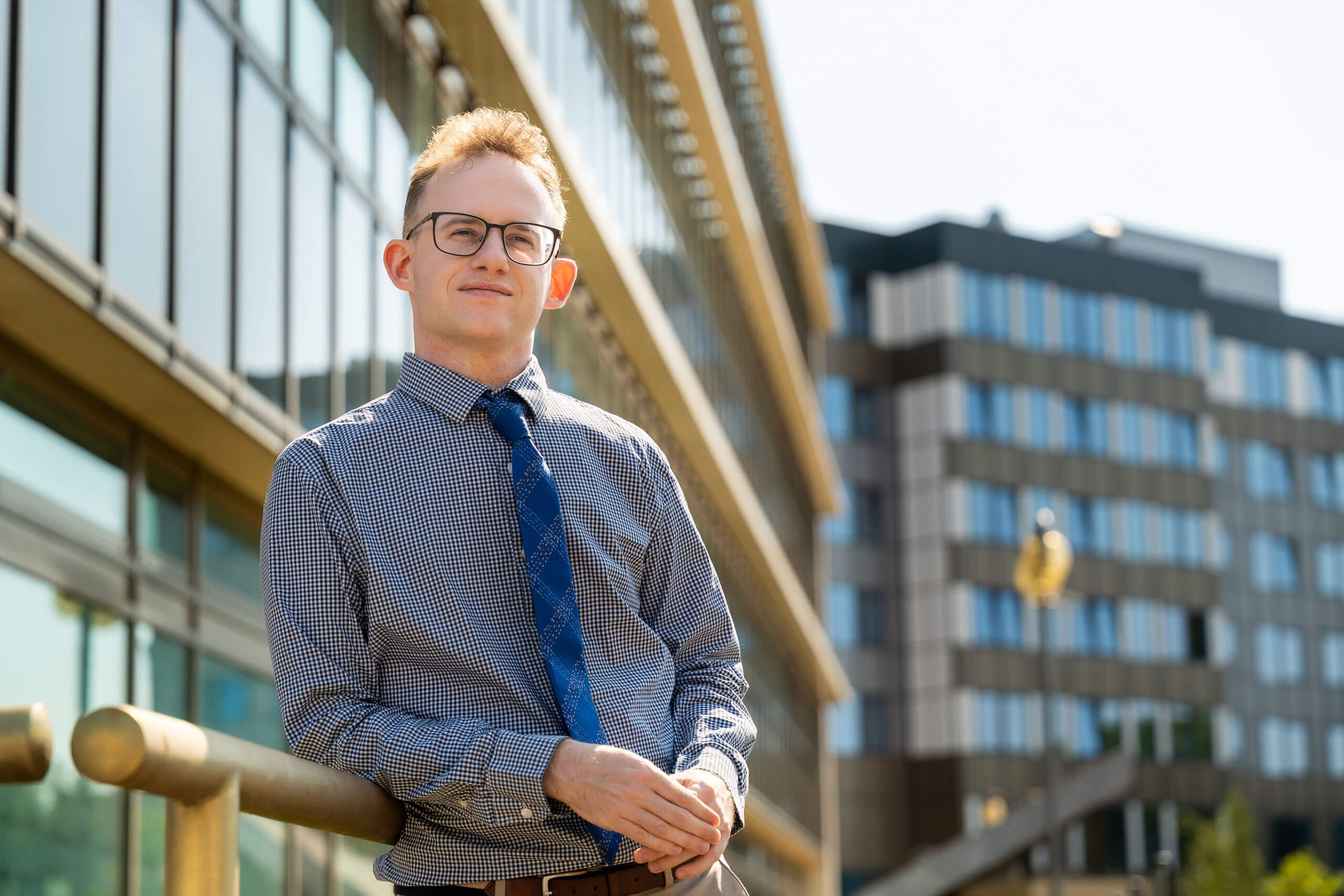 Piotr Alster, PhD hab., a young man wearing glasses, dressed in a shirt and tie, poses for a photo in front of buildings with glass windows. He smiles.