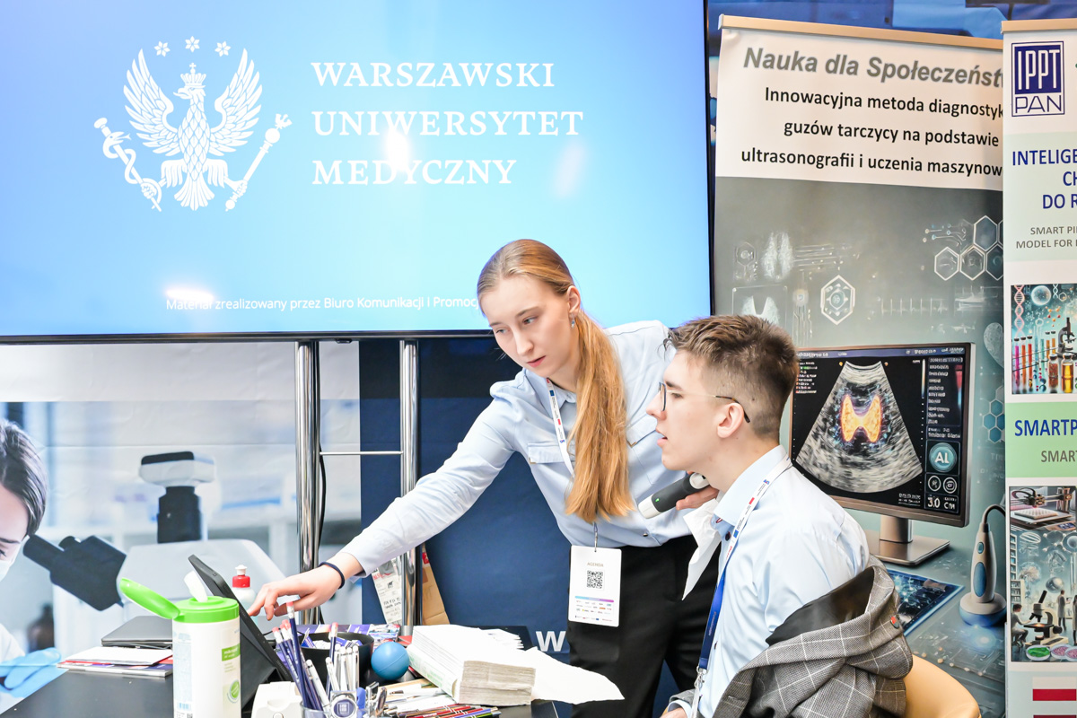 Two people are sitting at a desk in an office. A woman with long blonde hair is pointing at a computer screen. A man wearing glasses is looking at the screen. They are both wearing blue shirts and ID badges. There are tissues, pens, and a bottle of disinfectant on the desk. In the background, there is a large monitor and a poster with a medical image.