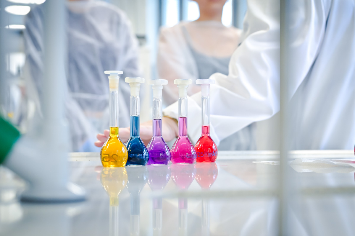 Na blacie w laboratorium stoi pięć kolb miarowych w rzędzie. Kolby mają różne kolory cieczy: żółty, niebieski, fioletowy, różowy i czerwony. W tle widać dwie osoby w białych fartuchach, jedna z nich ma rękę przy kolbach. W tle widać fragmenty sprzętu laboratoryjnego.