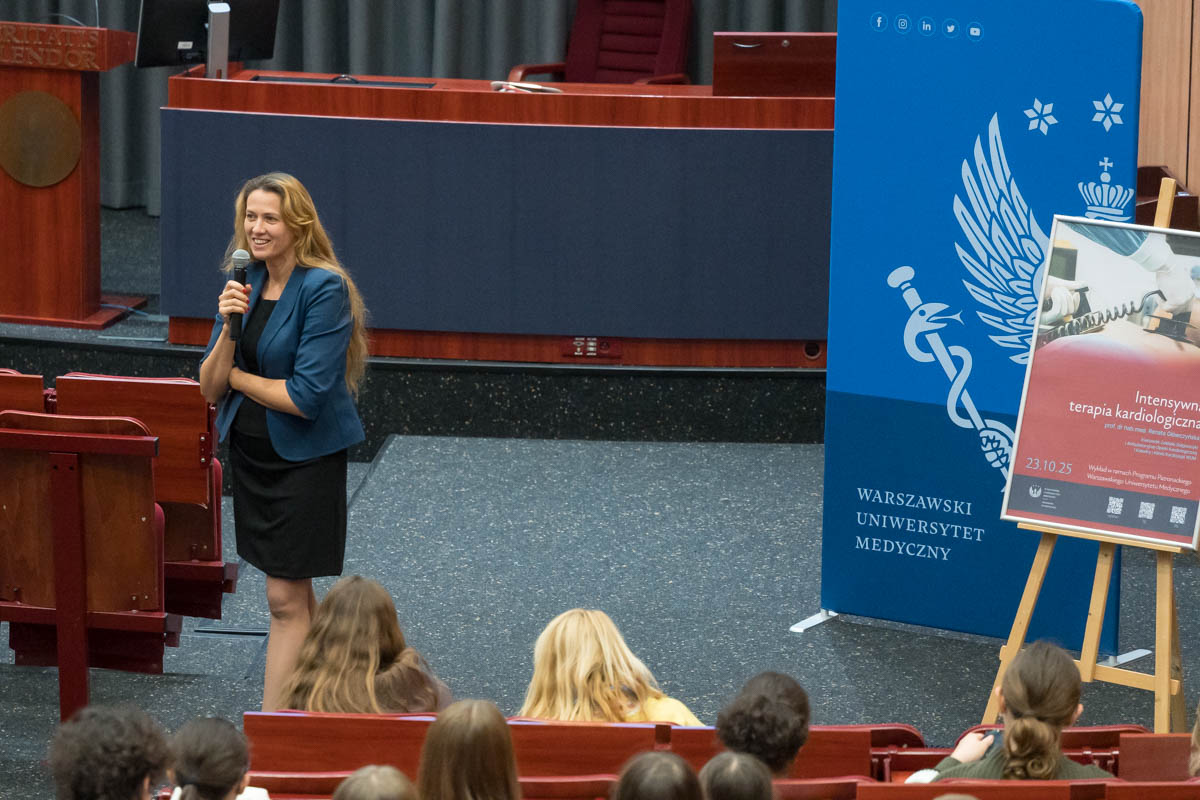 A person with a microphone is speaking in a lecture hall. Next to them is a blue banner reading “Warsaw Medical University” and a poster for a cardiology event. The audience is seated in red armchairs.