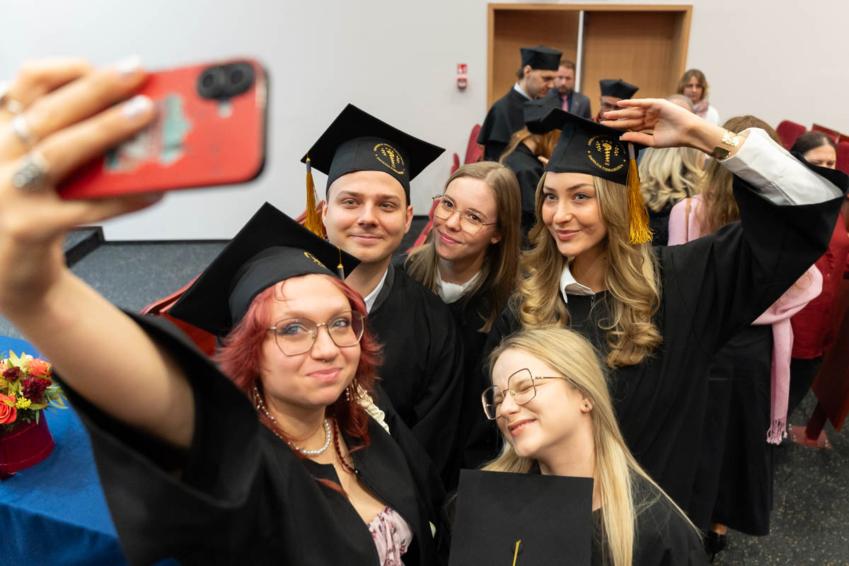 Sześć osób w togach i czapkach absolwenckich. Stoją blisko siebie w sali z czerwonymi krzesłami i drewnianymi drzwiami w tle. Jedna osoba trzyma telefon i robi selfie. Osoby pozują do zdjęcia, część unosi rękę w geście pozowania.