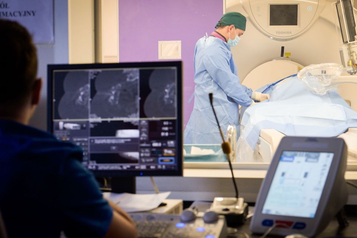 Osoba w niebieskim fartuchu ochronnym i zielonym czepku medycznym stoi przy stole zabiegowym, przykrytym sterylnym niebieskim materiałem, obok dużego aparatu diagnostycznego typu tomograf komputerowy. 