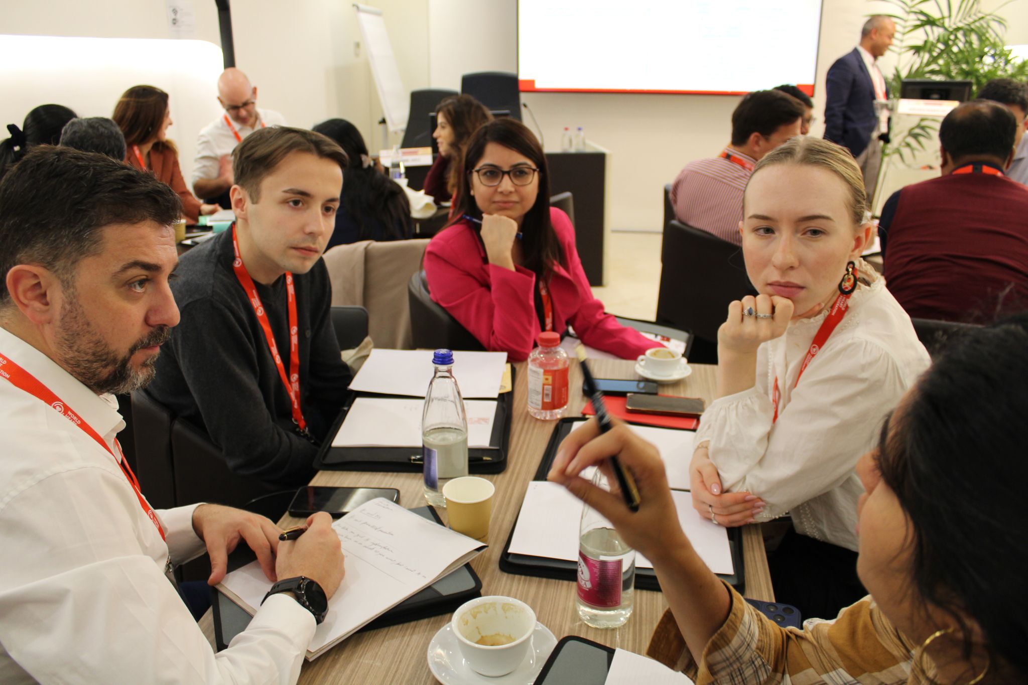 Przy stole siedzi sześć osób, mają przed sobą notatniki, długopisy, kubki z napojami i butelki wody. Na stole leży także tablet i telefon. W tle widać inne osoby przy stołach.