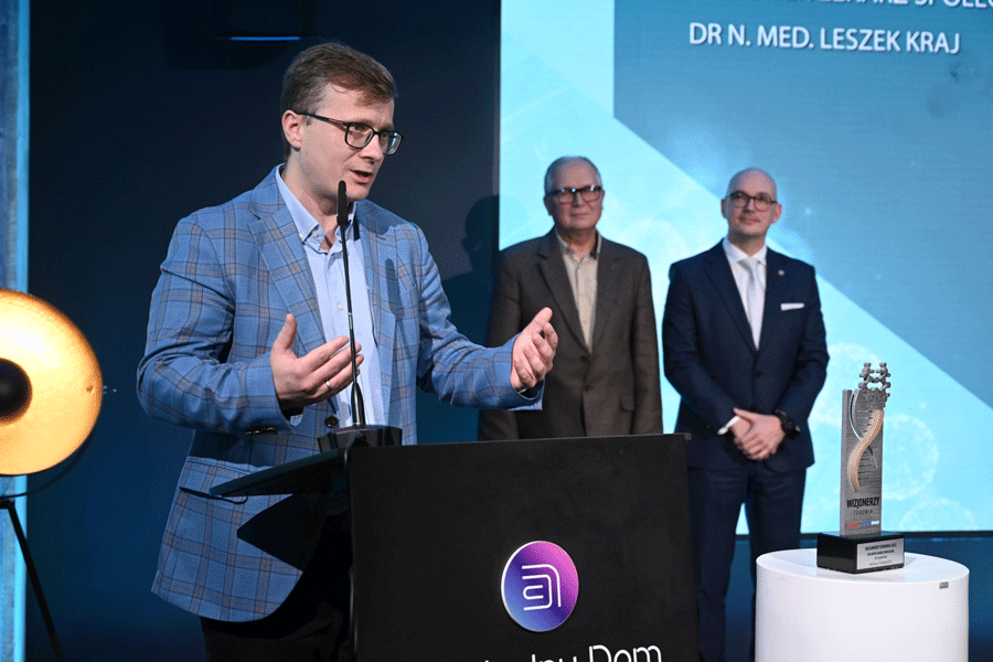 Three people standing on stage during an award ceremony. In the foreground is Dr. Leszek Kraj, standing behind a lectern and giving a speech. His hands are raised to chest height.
