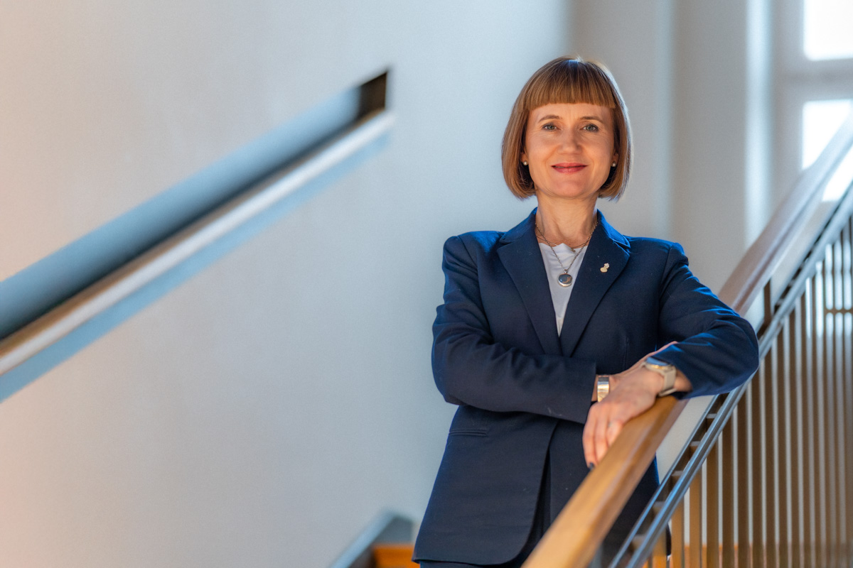 Kobieta w granatowej marynarce stoi na schodach oparta o poręcz.