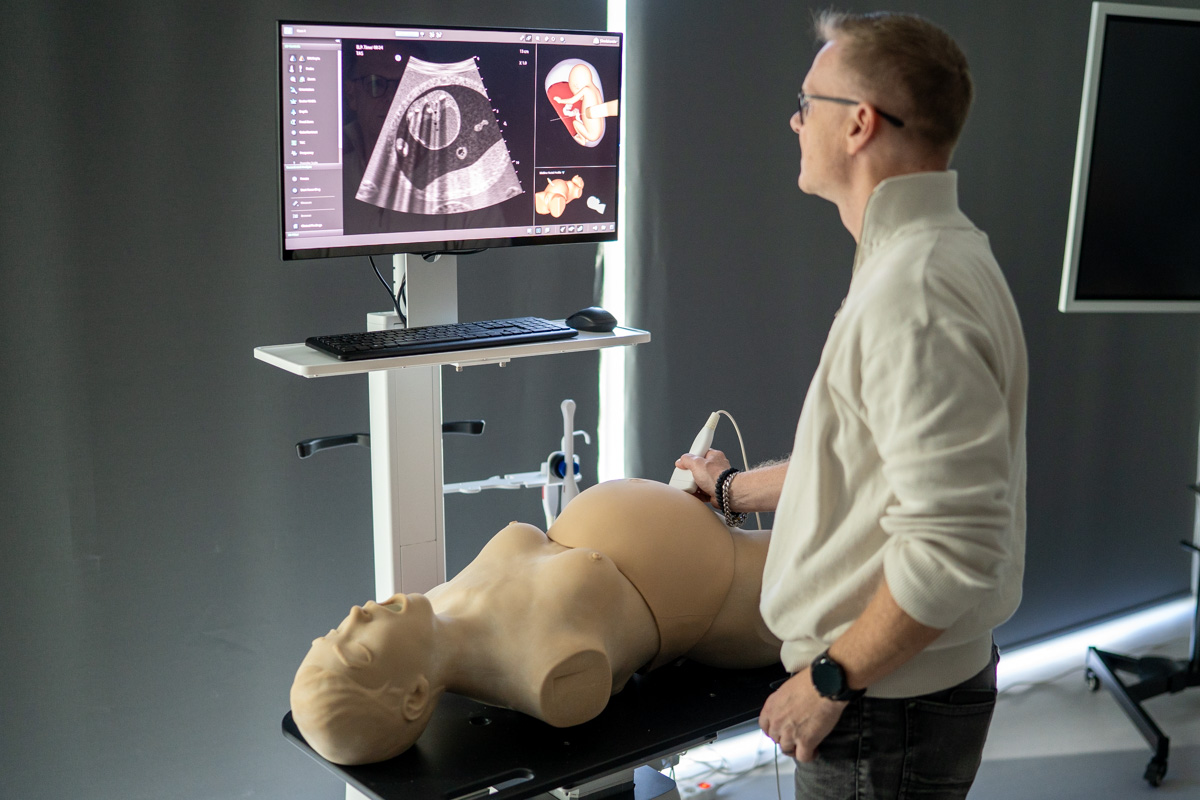 A man is standing next to a table with a realistic medical phantom on it. He's holding an ultrasound transducer in one hand and simulating an ultrasound exam. There's a monitor on a stand in front of him.