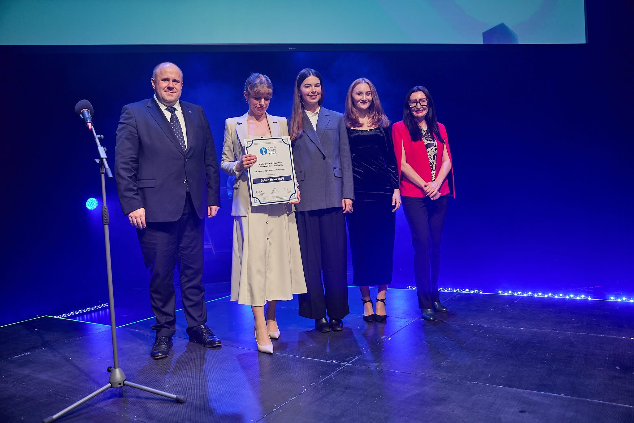 Five people are standing on stage in elegant attire. One person, the supervisor of the Gynecological Endoscopy Student Research Group, is holding a large frame with a certificate. 