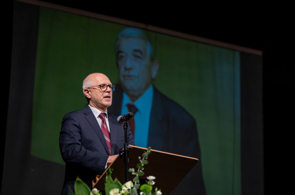 The rector, Prof. Rafał Krenke, elegantly dressed, stands behind the lectern and gives a speech. In the background, there is a photo of an elderly man—Prof. Zbigniew Religa.