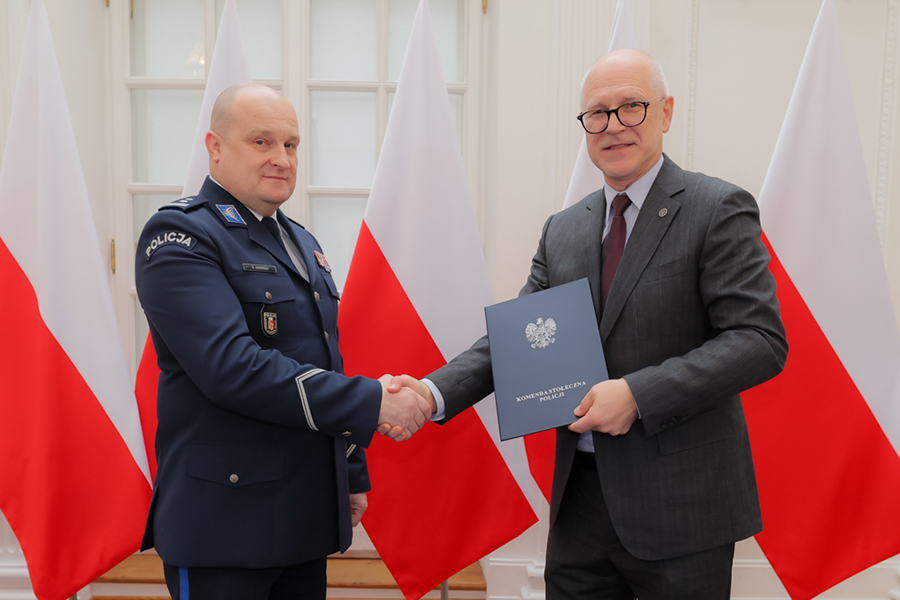 Dwóch mężczyzn, ten po lewej ubrany w mundur policyjny, ten po prawej - w garnitur, ściskają sobie dłonie. Mężczyzna po prawej trzyma w ręce zamkniętą teczkę. W tle widać flagi RP.