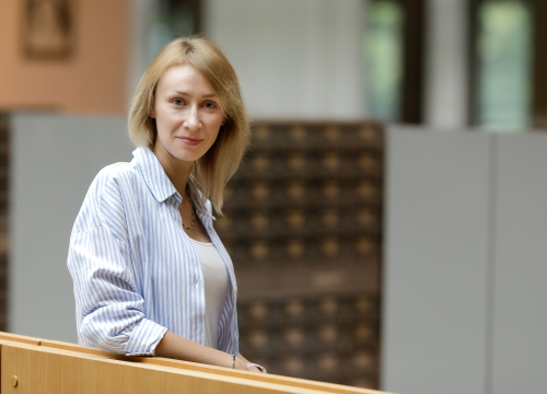 Młoda kobieta z jasnymi włosami opiera się o drewnianą balustradę. Uśmiecha się do aparatu. W tle regały biblioteczne.